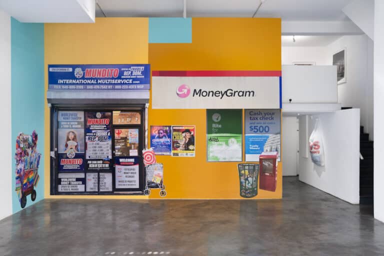 A street vendor’s cart and a hand-held cart stand in front of a colorful wall displaying ads for wire transfer services and concert posters.