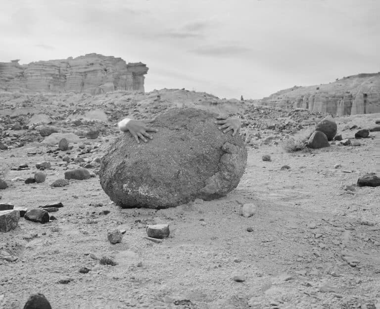 At the center of a rocky, desert landscape two hands appear from behind a large stone as if to embrace it.
