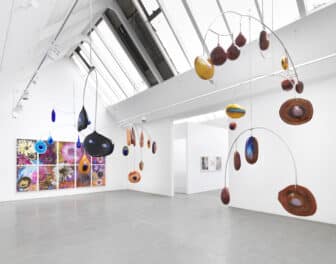 Round glass forms are suspended from a series of mobiles in a gallery. The forms vary in color and shape, including teal, blue, brown, black, and orange with colorful accents.