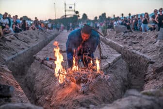 A man kneels in front of a fire conducting a performance piece while spectators on either side look on.