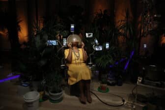 A person with dark skin wearing a yellow dress is sitting and pulls an opaque plastic helmet over their face. They are surrounded by small video screens and tropical foliage.