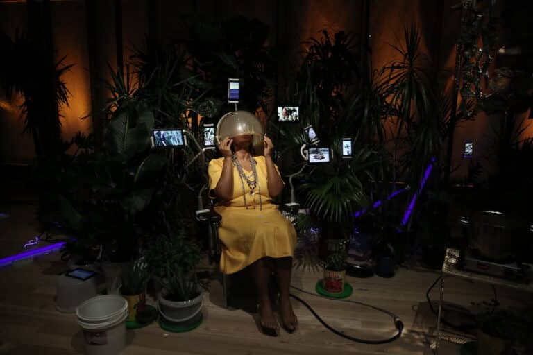 A person with dark skin wearing a yellow dress is sitting and pulls an opaque plastic helmet over their face. They are surrounded by small video screens and tropical foliage.