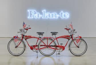 Two red Schwinn bicycles bearing American and Puerto Rican flags are connected by a shared back wheel and face opposing directions. They are positioned in front of a pale blue neon sign reading “Pa-lan-te”.