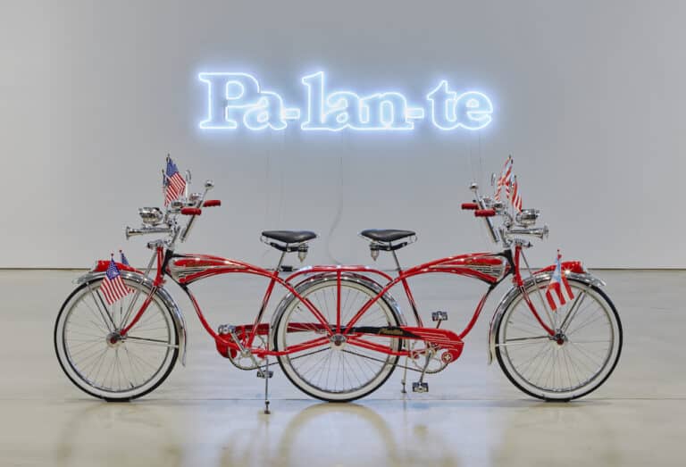 Two red Schwinn bicycles bearing American and Puerto Rican flags are connected by a shared back wheel and face opposing directions. They are positioned in front of a pale blue neon sign reading “Pa-lan-te”.
