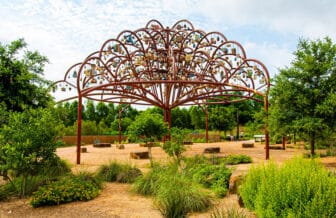 A large metal sculpture resembling a tree stands within a park. From the top of the tree hang seven hundred small sculptures representing the stories of community members.