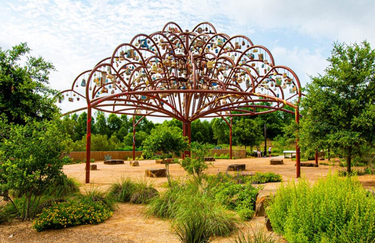 A large metal sculpture resembling a tree stands within a park. From the top of the tree hang seven hundred small sculptures representing the stories of community members.