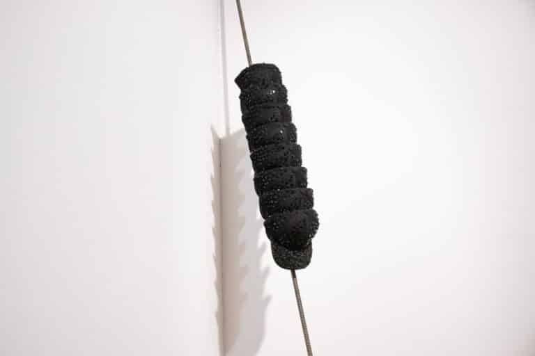 Seven black baseball caps covered in shattered polarized glass are stacked on each other and hung vertically on a steel rod.
