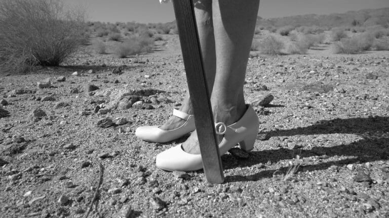 A film still in black and white shows two feet in a pair of low-heeled white shoes against a desert landscape.