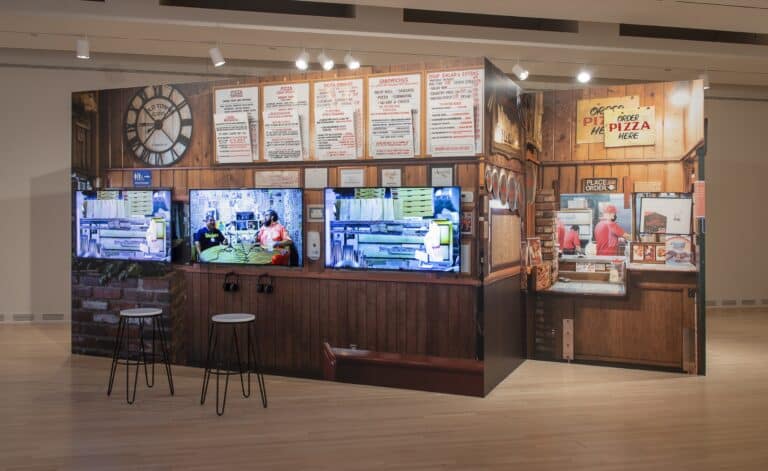 A structure resembling a restaurant that serves pizza is installed in a gallery, including menus and a register. On the walls of the installation are video screens.
