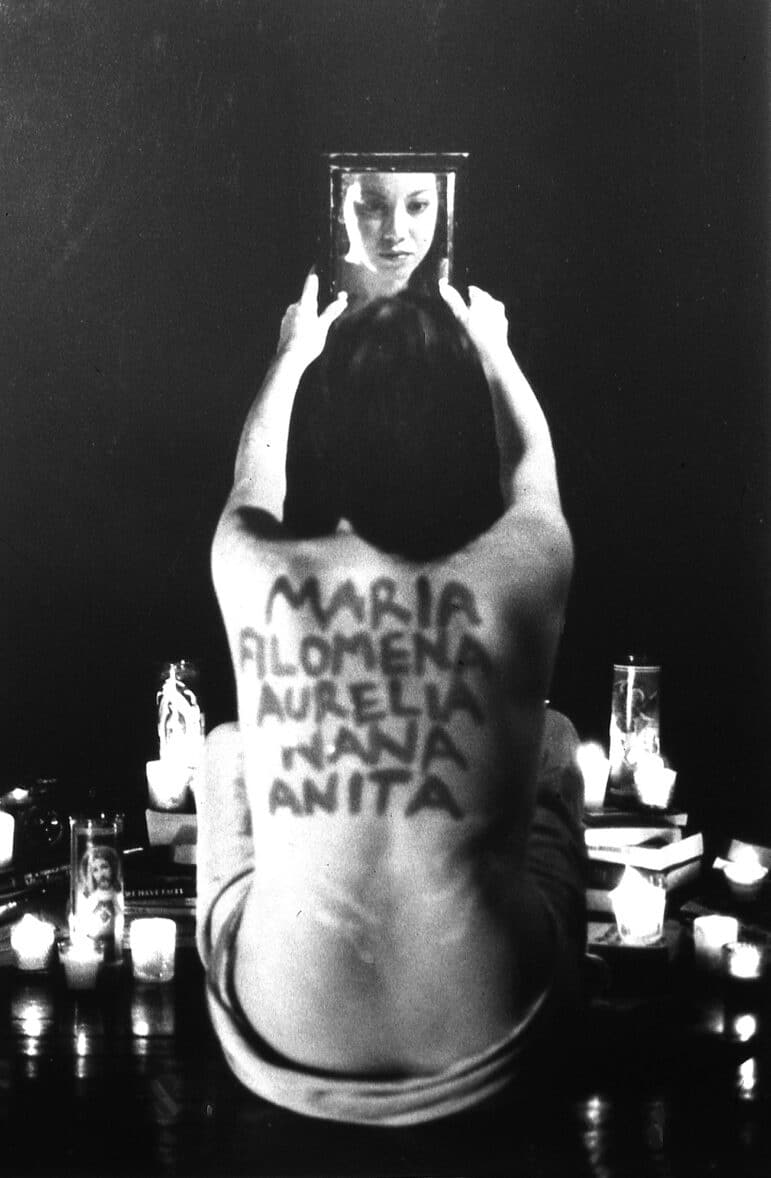 A self-portrait, previously also titled “Christina: Self Portrait,” that includes the artist’s back, inscribed with her female-identified family members’ names, her face reflected in a mirror that she holds up, and votive candles surrounding her person.