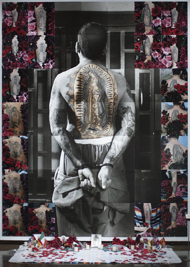 In a wall-sized photographic collage, a prisoner, identified by his jumpsuit and handcuffs, stands facing away from the camera, in front of a set of iron bars. The jumpsuit is rolled down to his waist so as to expose his naked back, on which is emblazoned a large color tattoo of Our Lady of Guadalupe. Around the perimeter of the image are small color photographs of various other prison tattoos of Our Lady of Guadalupe on other men.