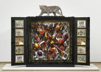 A jaguar statue sits atop a tri-fold display covered in black fur with framed black and white images to the right and left of a middle section that includes an assortment of books, human remains, and other objects dipped in blood and gold.