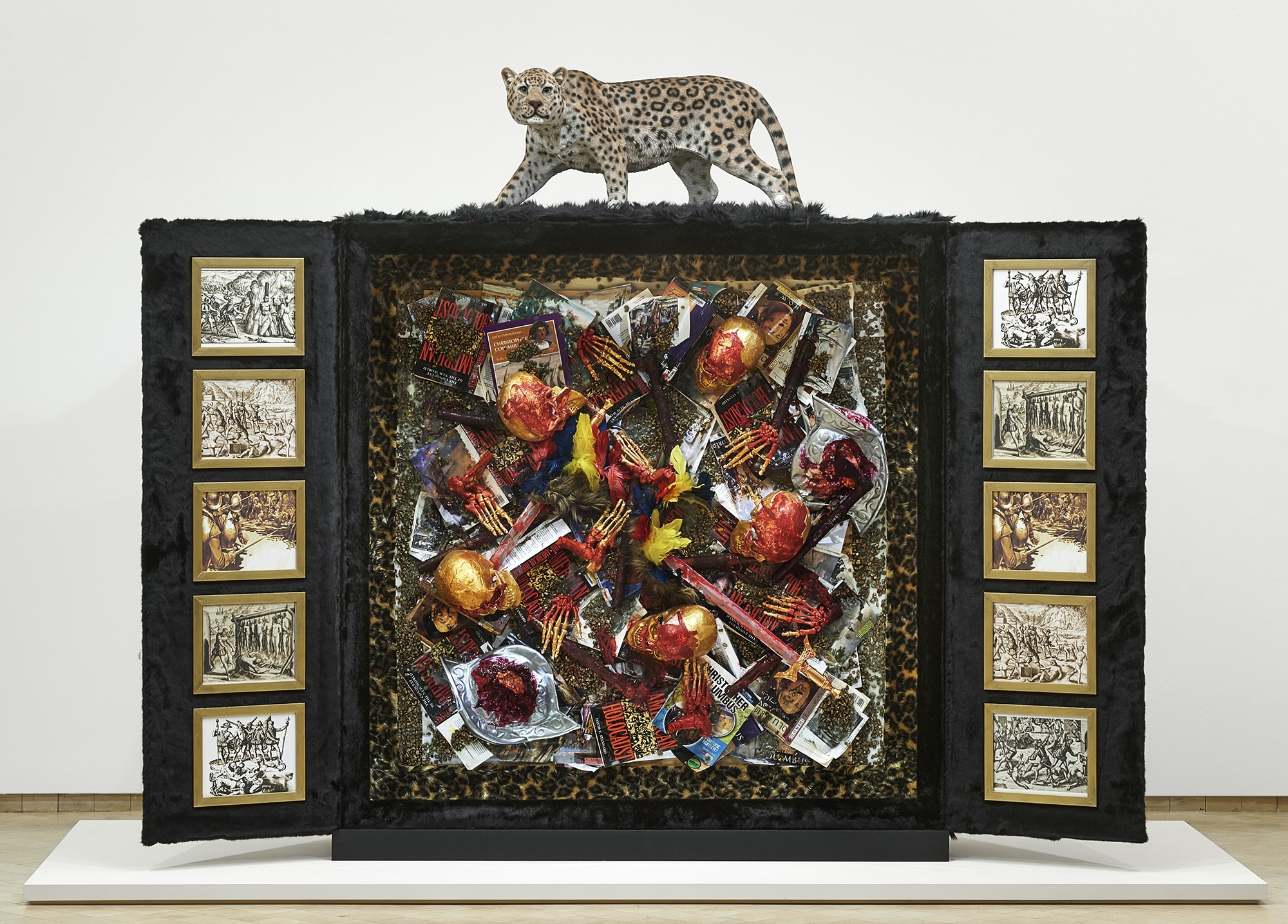 A jaguar statue sits atop a tri-fold display covered in black fur with framed black and white images to the right and left of a middle section that includes an assortment of books, human remains, and other objects dipped in blood and gold.