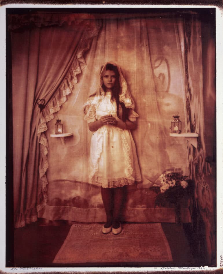 A preteen girl dressed in a white first communion gown and lace-trimmed veil stares directly into the camera, her hands clasped around a rosary in front of her. She is standing on a small rug and is framed from behind by several curtains, with small shelves that hold lit candles on either side of her.
