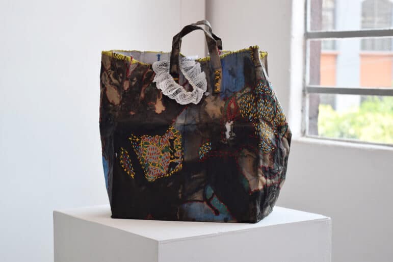 A large paper shopping bag displayed on a white pedestal in a gallery setting with a window visible in the background. The bag’s surface is heavily worked with dark, layered washes of ink and watercolor in deep blues, browns, and blacks. Scattered across the surface are areas of dense embroidery in bright yellows, reds, and greens, with visible stitching that patches and mends sections of the bag. A white lace ruffle is attached near one of the handles at the top. The bag retains its functional shape but bears signs of repair and embellishment, giving it the quality of both a worn object and a finished artwork.