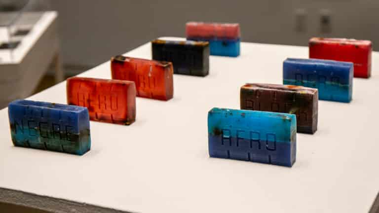 Nine rectangular handmade bars of soap arranged in two rows on a white display surface in a gallery setting. Each bar is composed of layered colors, primarily deep blues, bright turquoises, reds, and dark browns, giving them a striated, geological appearance. Each bar is embossed with a word pressed into its surface; visible words include “NEGRE,” “AFRO,” and “BLACK.” The bars vary slightly in color combination and show traces of organic material embedded within the glycerin. Their handmade quality is evident in the slight irregularities of their surfaces and the uneven distribution of color and herbs throughout each bar.