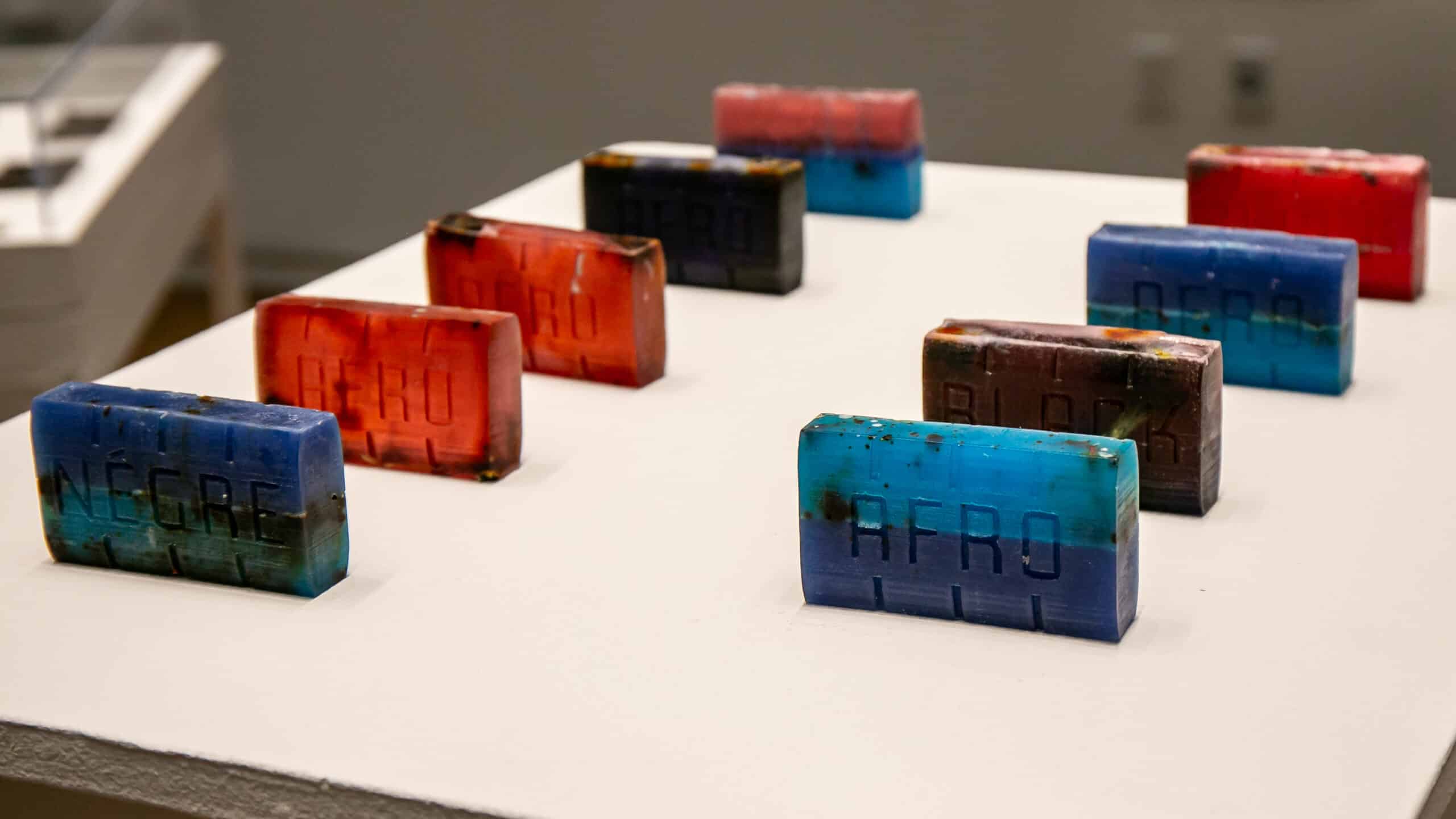 Nine rectangular handmade bars of soap arranged in two rows on a white display surface in a gallery setting. Each bar is composed of layered colors, primarily deep blues, bright turquoises, reds, and dark browns, giving them a striated, geological appearance. Each bar is embossed with a word pressed into its surface; visible words include “NEGRE,” “AFRO,” and “BLACK.” The bars vary slightly in color combination and show traces of organic material embedded within the glycerin. Their handmade quality is evident in the slight irregularities of their surfaces and the uneven distribution of color and herbs throughout each bar.