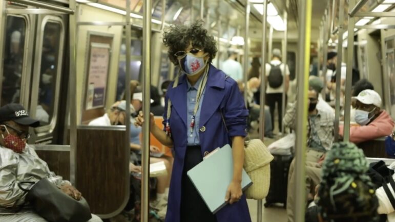 A still from a video: the artist, in character as an investigator, stands at the center of a crowded subway car, gripping a vertical pole with one hand and clutching a pale-blue folder under the other arm. She wears a blue trench coat, round sunglasses, and a printed face mask, with a woven bag on her shoulder; masked passengers sit and stand around her.