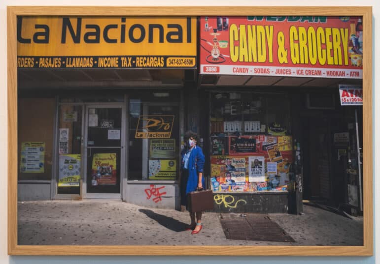 A framed color photograph of the artist in character as an investigator—blue coat, printed face mask, red heels, brown briefcase—standing on a sunlit sidewalk between two Dominican-owned storefronts, a “La Nacional: money-transfer office and a red “Candy & Grocery” bodega.
