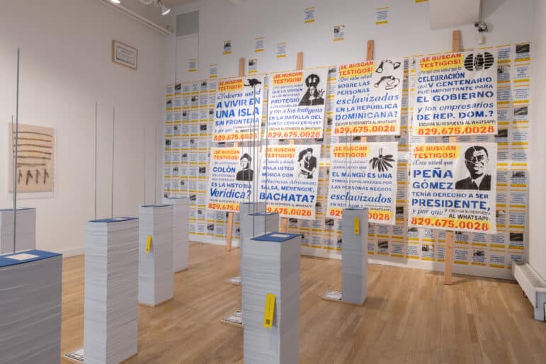 A gallery installation view with walls covered in screen-printed banners and flyers reading “¡Se buscan testigos!” (Looking for Witnesses!), each posing a question in Spanish about Dominican history and race above a WhatsApp number; tall columns of stacked paper stand on raw wooden posts in the foreground.