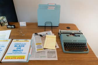 A wooden desk staged as a detective’s workstation: a vintage teal typewriter at right, a stand of pale-blue date-labeled file folders at center, and across the surface a yellow legal pad of handwritten notes, newspapers, round sunglasses, business cards printed with a WhatsApp number, and an open folder showing a flyer that reads “Looking for Witnesses! Dominican, do you consider yourself Black or Indio? Did migrating change your perspective?”