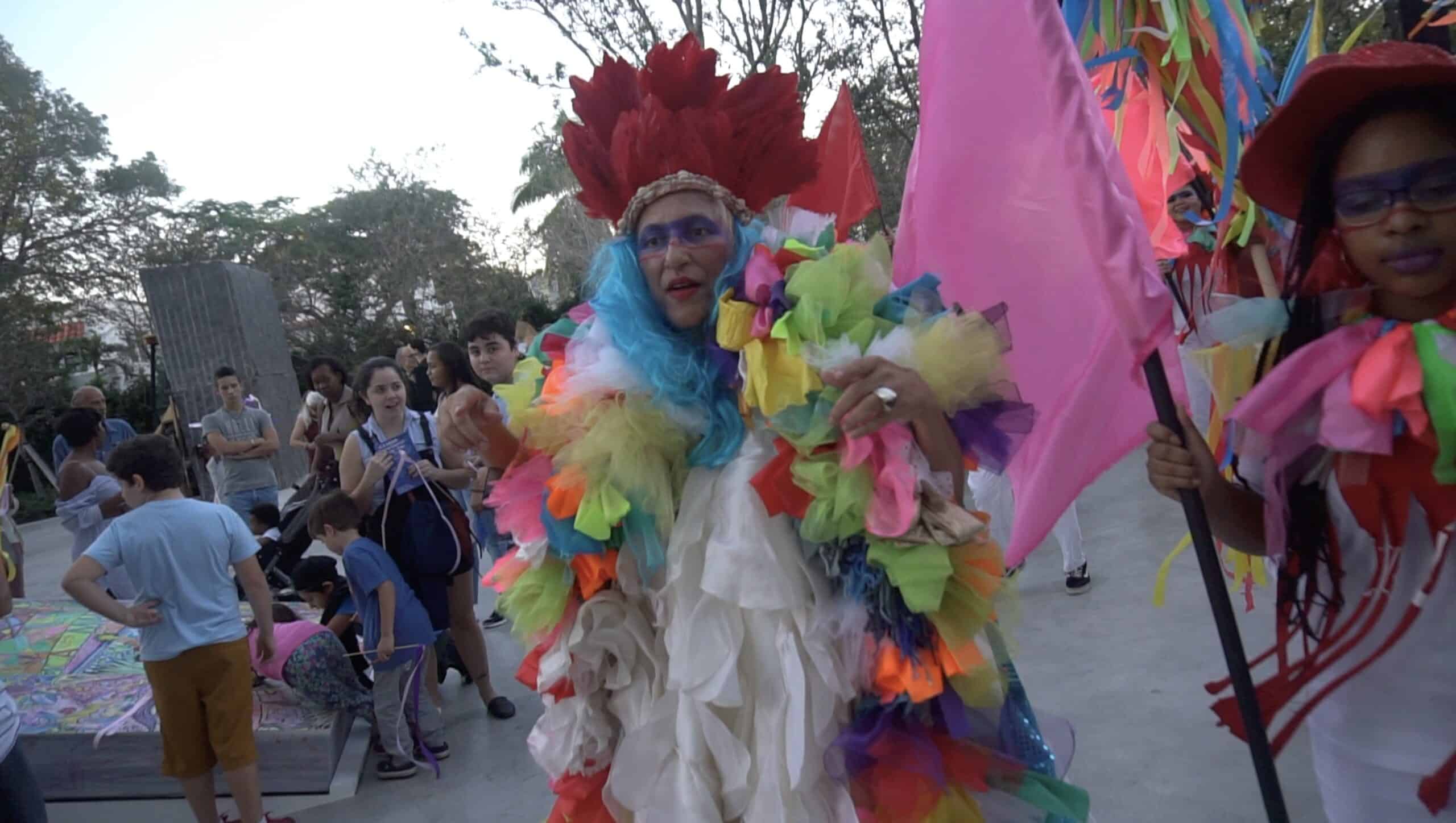 Charo Oquet leads a colorful Rará-inspired procession, wearing a vibrant costume of multicolored fabric and a feathered headdress while carrying a pink flag. Community members gather around her, joining the festive performance through music, movement, and shared participation.