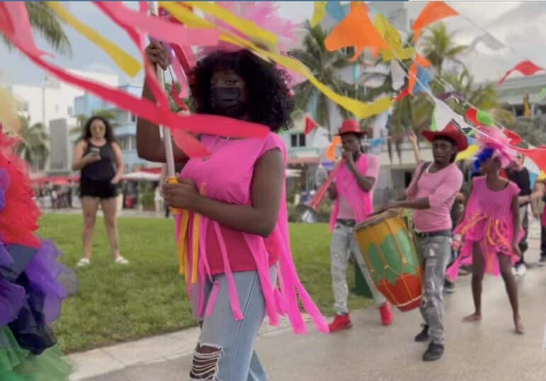 Participants in a Rará-inspired procession carry colorful streamers and play drums and horns as they move through a public park. Bright pink costumes, ribbons, and music transform the outdoor setting into a festive, communal performance.