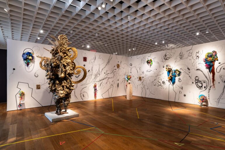 Installation view of Charo Oquet’s “I Am Here: Fragments, Trances, and Improvised Architectures,” featuring a monumental bronze-toned sculpture surrounded by colorful assemblages and expressive wall drawings. The immersive gallery evokes African diasporic spiritual traditions through layered materials, symbolic forms, and a dynamic sense of transformation and connection.