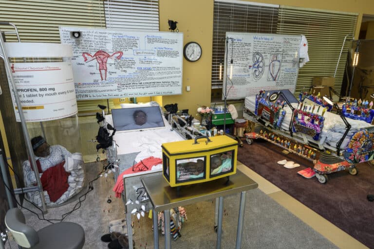Two hospital beds form part of an art installation. A video monitor showing a patient telling their story rests on each bed. Behind the bed, a whiteboard displays handwritten notes about each patient’s illness.