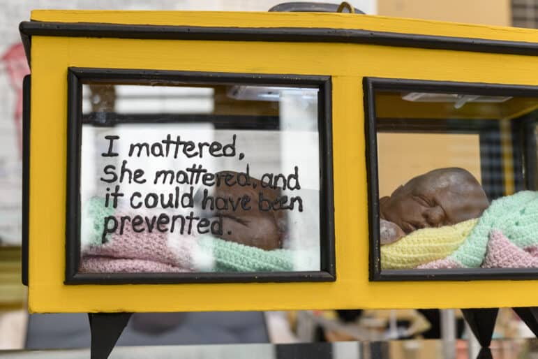 A sculpture of an infant is visible inside a small yellow display case. On the outside of the case is handwritten: “I mattered, she mattered, and it could have been prevented.”