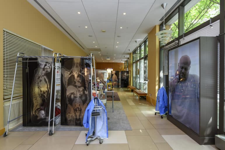 A large room is separated into spaces for hospital beds by room dividers made from X-ray prints. A tall video monitor shows a man in a hospital gown grimacing in pain.