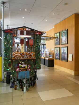 In a large room, four photographic portraits of healthcare workers hang on a wall. A freestanding sculpture made from a mobile food cart is embellished with pill bottles, hot-water bottles, and other items.