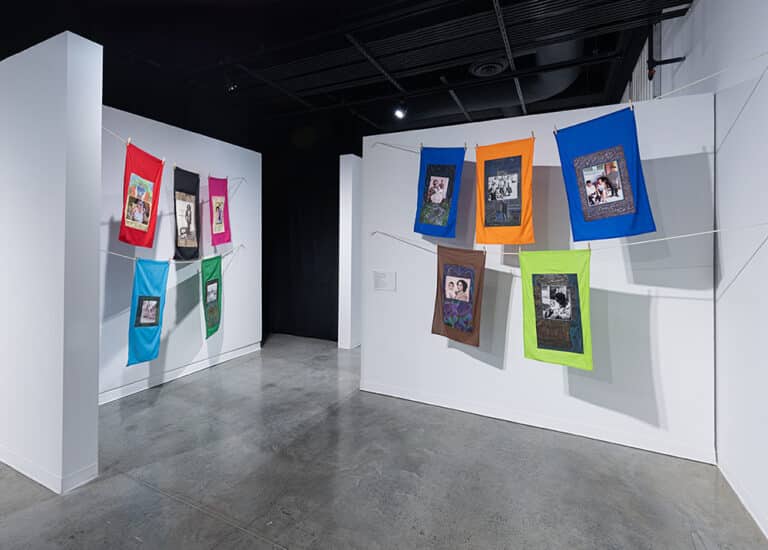 Ten brightly colored textile works hung with clothespins on stacked clotheslines that form four rows across two adjacent walls.