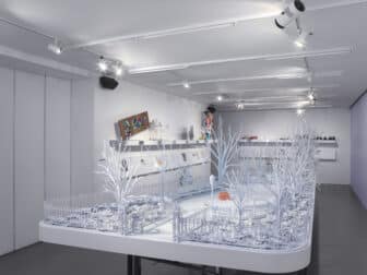 An all-white architectural maquette of a city park stands on a platform in a gallery with white walls and shelves with colorful sculptures visible in the background.