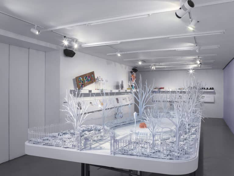 An all-white architectural maquette of a city park stands on a platform in a gallery with white walls and shelves with colorful sculptures visible in the background.