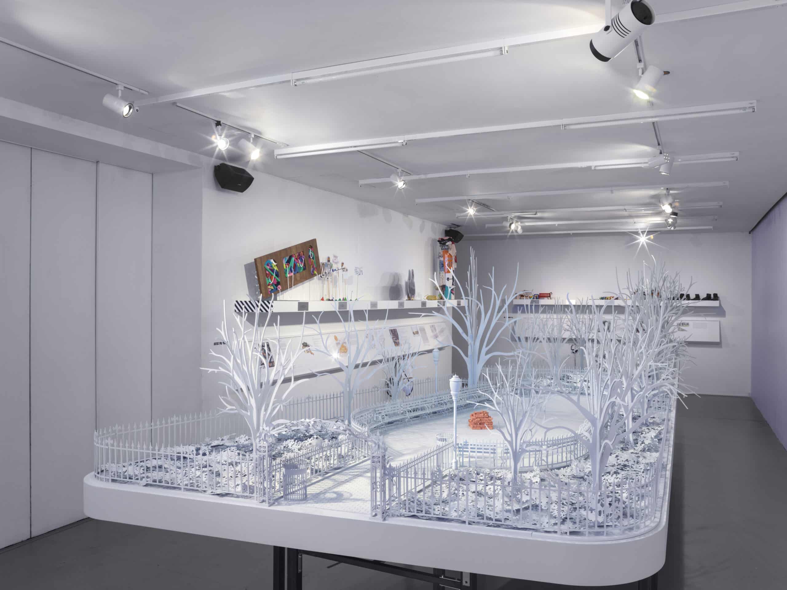 An all-white architectural maquette of a city park stands on a platform in a gallery with white walls and shelves with colorful sculptures visible in the background.