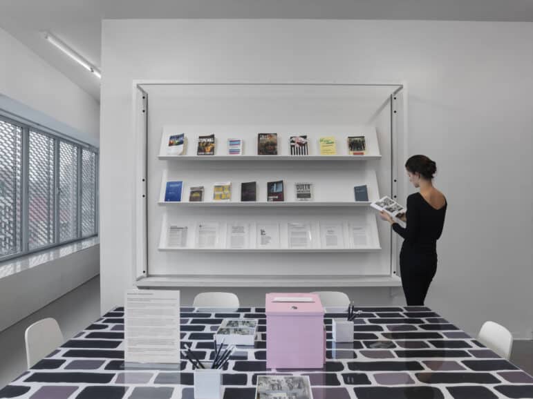 In the foreground, a table with a small white standing plexiglass sign with small text, and a pink suggestion box. In the background, a row of three white wall-mounted shelves displaying book publications. A woman with dark hair wearing a close-fitting back outfit holds a book open, reading.