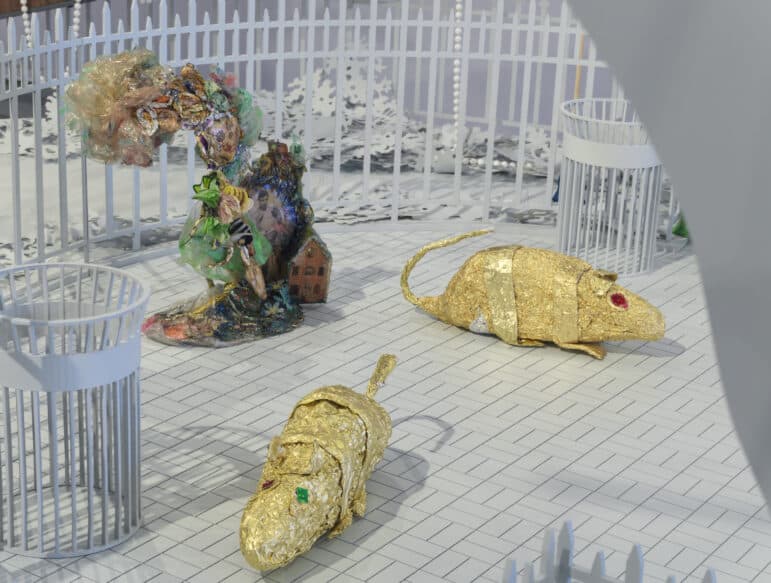 Within the all-white architectural maquette of a city park are two rats made of gold foil with red and green eyes drawn on with marker. Behind them is a miniature tower made of crumpled foil and cellophane. Embedded within the tower is a black-and-white photo of a man, his fists held up, and a model of a house.