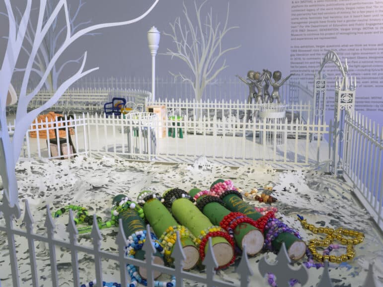 Within the all-white architectural maquette of a city park are five cross-cut wooden cylinders wrapped with green paper and wrapped on each end with bright plastic Mardi Gras beads.
