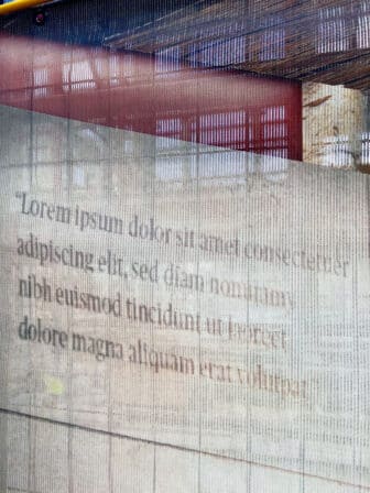 A detailed view of a semi-transparent windscreen with a paragraph of lorem ipsum text layered over a thick-gauge steel wire fence with rectangular panels. The placeholder text reads, “Lorem ipsum dolor sit amet, consectetur adipiscing elit, sed do eiusmod tempor incididunt ut labore et dolore magna aliquam erat voluptat.”