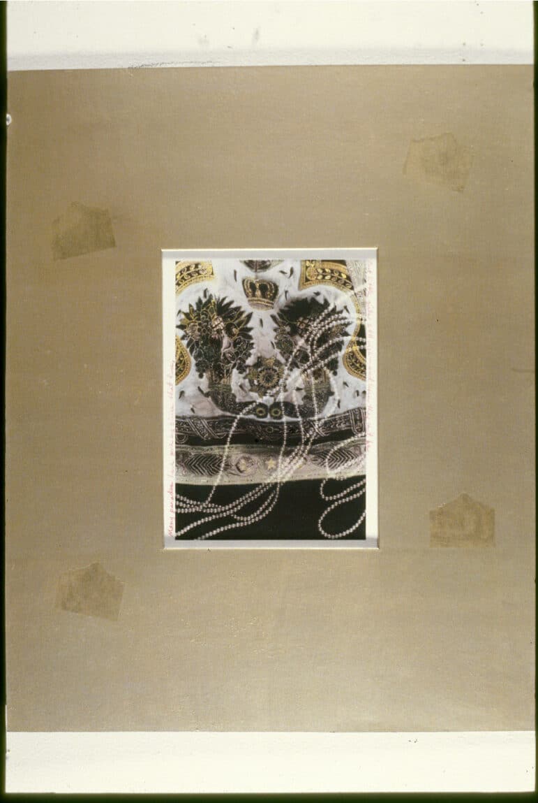 A close-up of a heavily embellished textile with a ceremonial crown and ornamental motifs tinted a metallic gold. Small strings of pearls are placed horizontally along the right side, accompanied by two sentences written in red pencil along the white edges.