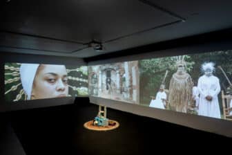 This image shows the installation of “Babalú-Ayé y La Negra del Hospital” (2022) at the Fridman Gallery in New York. In a dark room, there are three large panoramic projections on adjacent walls surrounding a small sculpture composed of cinder blocks decorated with cowrie shells placed in the off-center right side of the gallery. The video, still on the left side, shows a Black woman with a white headpiece with cowrie shells looking directly at the viewer. In the center there is a figure dancing in the middle of colonial architecture structures. The image to the far right shows three figures dressed in ceremonial attires that display Black diasporic culture. One of the figures is sitting on a rocking chair while the other two people stand facing forward.