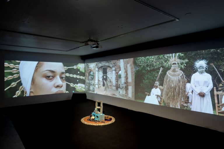 This image shows the installation of “Babalú-Ayé y La Negra del Hospital” (2022) at the Fridman Gallery in New York. In a dark room, there are three large panoramic projections on adjacent walls surrounding a small sculpture composed of cinder blocks decorated with cowrie shells placed in the off-center right side of the gallery. The video, still on the left side, shows a Black woman with a white headpiece with cowrie shells looking directly at the viewer. In the center there is a figure dancing in the middle of colonial architecture structures. The image to the far right shows three figures dressed in ceremonial attires that display Black diasporic culture. One of the figures is sitting on a rocking chair while the other two people stand facing forward.