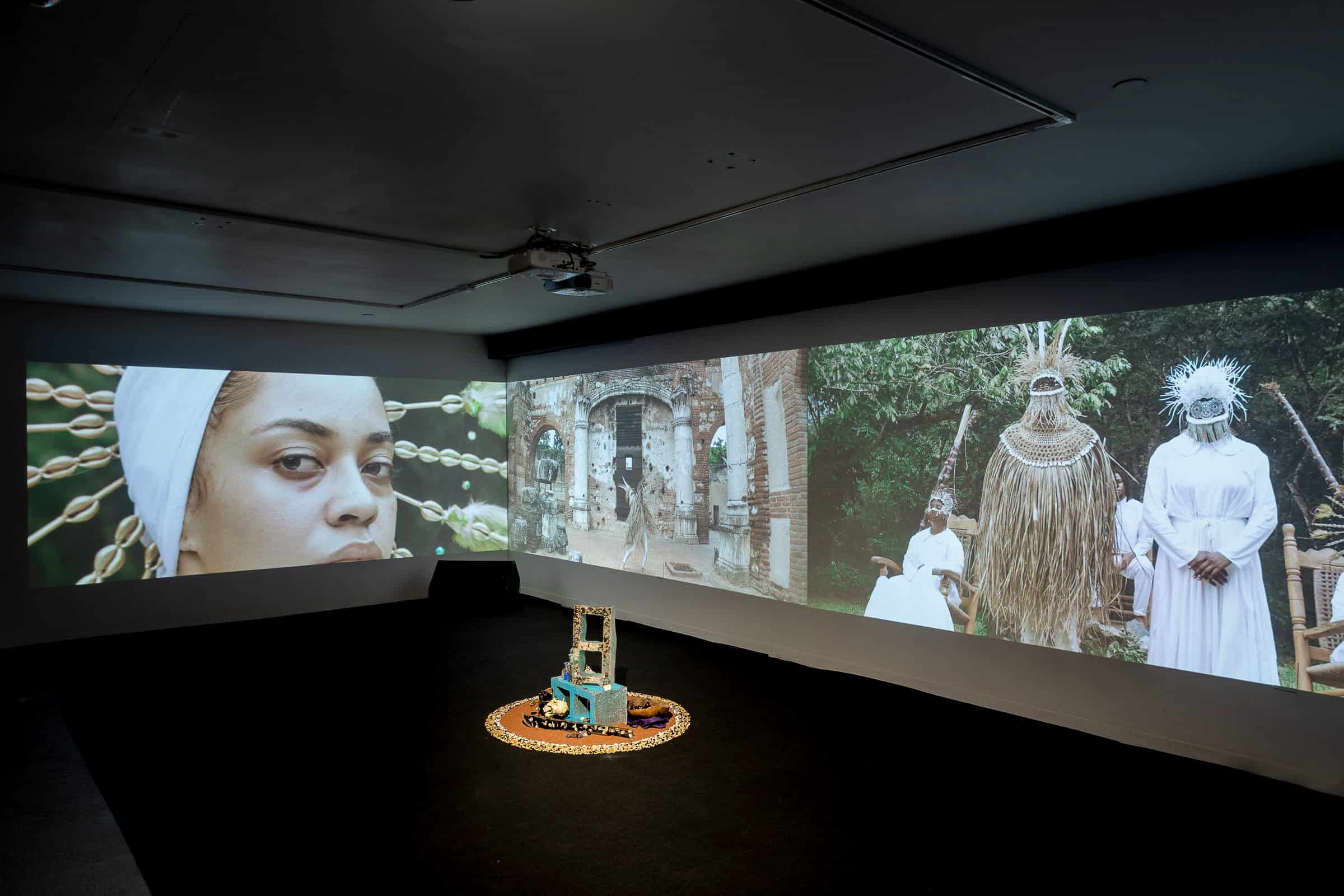 This image shows the installation of “Babalú-Ayé y La Negra del Hospital” (2022) at the Fridman Gallery in New York. In a dark room, there are three large panoramic projections on adjacent walls surrounding a small sculpture composed of cinder blocks decorated with cowrie shells placed in the off-center right side of the gallery. The video, still on the left side, shows a Black woman with a white headpiece with cowrie shells looking directly at the viewer. In the center there is a figure dancing in the middle of colonial architecture structures. The image to the far right shows three figures dressed in ceremonial attires that display Black diasporic culture. One of the figures is sitting on a rocking chair while the other two people stand facing forward.